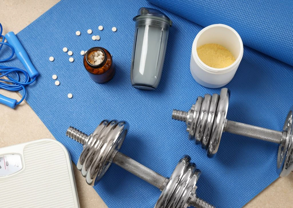 Dumbbells, protein shake and supplements on a yoga mat. 